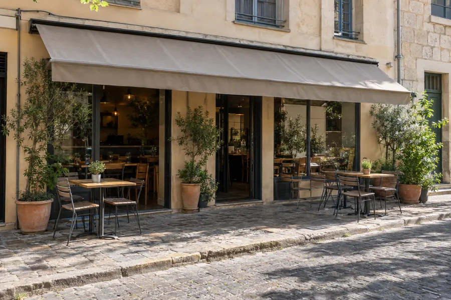 Un piccolo ristorante con dehors e una semplice tenda da sole, visto dall’altro lato di una strada tranquilla in una mattina di sole