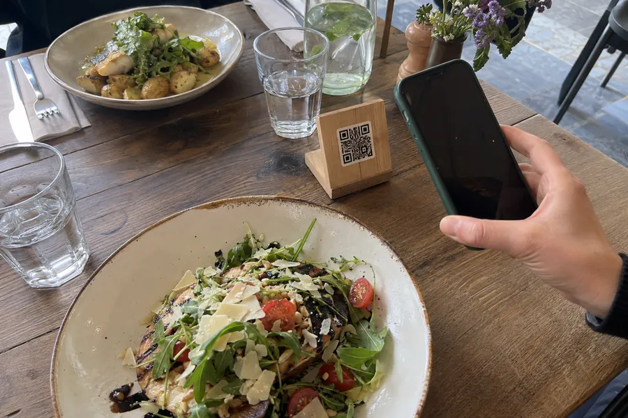A lunch table with two plates, glasses of water and a small QR code stand, a guest holding a phone next to it
