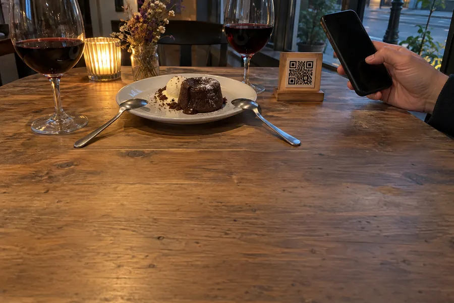 Ein Schokoladenfondant, zwei Gläser Wein und ein kleiner QR-Code-Aufsteller auf einem Bistrotisch, eine Hand hält ein Handy