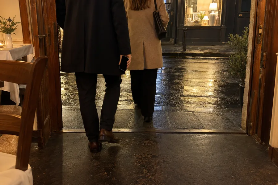 Clienti con il cappotto che escono da un piccolo bistrot a tarda sera, visti di spalle, uno con il telefono in mano; dalla sala filtra una luce calda