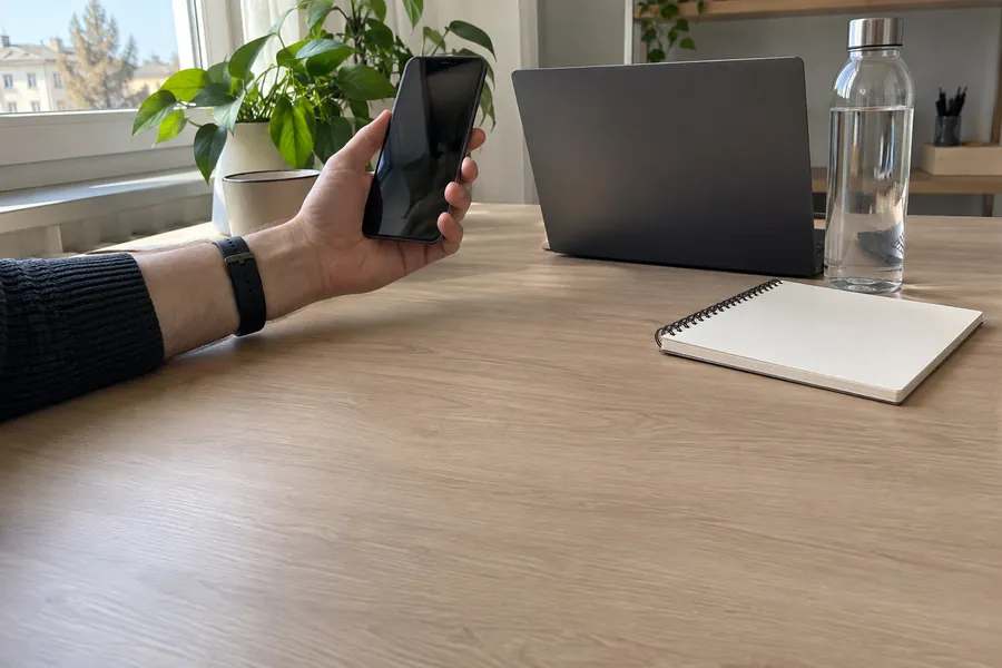 A hand holding a phone above an office desk with a laptop and a notebook, late morning