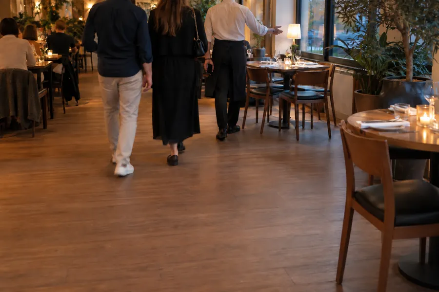 Un cameriere accompagna due clienti appena arrivati tra i tavoli di un bistrot pieno, la sera