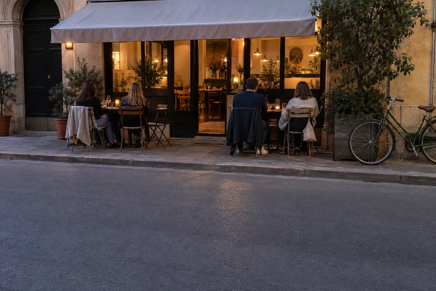 Un piccolo ristorante all’imbrunire visto dall’altro lato della strada, con una luce calda alle finestre e clienti nel dehors
